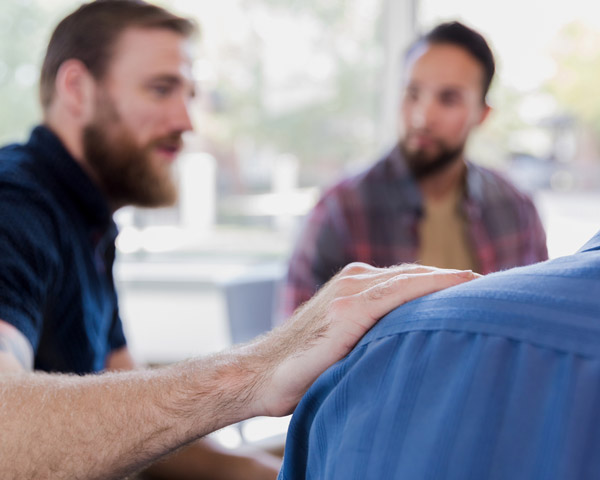 During a support group session a mid adult man comforts his friend. His hand is on the man's shoulder.