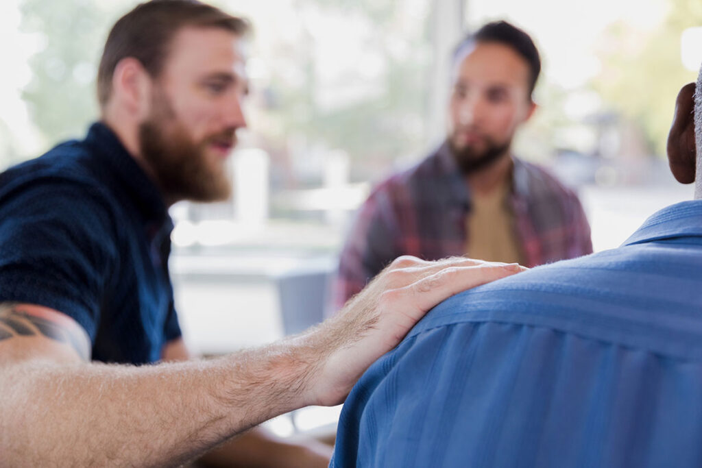 During a support group session a mid adult man comforts his friend. His hand is on the man's shoulder.